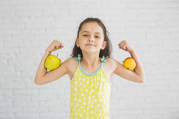 niña flexiona sus brazos, sosteniendo en sus biceps manzanas, lo que nos sugiere habitos saludables como alimentación y ejercicio